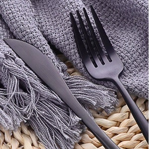 May include: Close-up of a matte black knife and fork set. The knife has a serrated edge, and the fork has four tines. The silverware is placed on a gray fringed cloth and a woven surface.