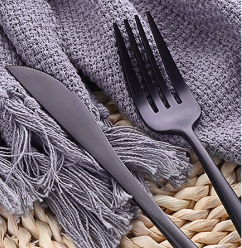 May include: Close-up of a matte black knife and fork set. The knife has a serrated edge, and the fork has four tines. The silverware is placed on a gray fringed cloth and a woven surface.