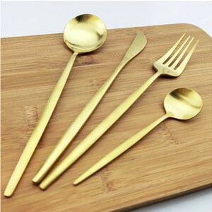 May include: A set of four gold-colored utensils: a soup spoon, a butter knife, a fork, and a smaller spoon. The utensils are arranged on a wooden cutting board, showcasing their sleek, modern design. The spoons have round bowls, and the fork has four tines.