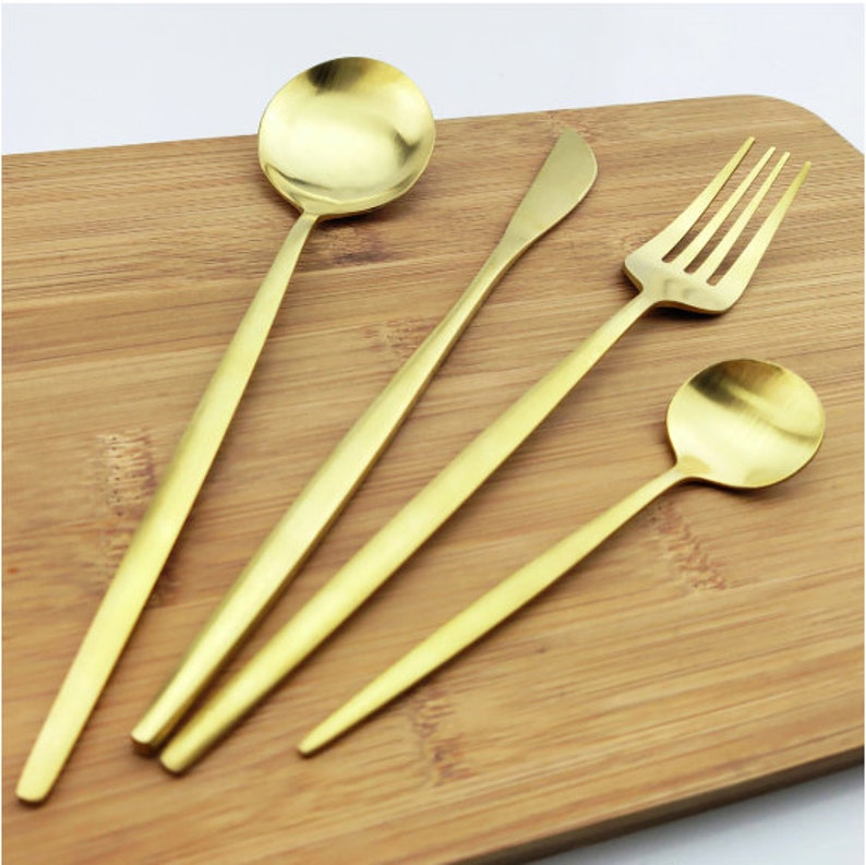 May include: A set of four gold-colored utensils: a soup spoon, a butter knife, a fork, and a smaller spoon. The utensils are arranged on a wooden cutting board, showcasing their sleek, modern design. The spoons have round bowls, and the fork has four tines.