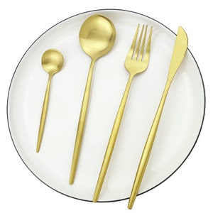 May include: A set of four gold-colored utensils: a small spoon, a large spoon, a fork, and a knife. The utensils are arranged on a white plate with a black rim. The utensils have a sleek, modern design.