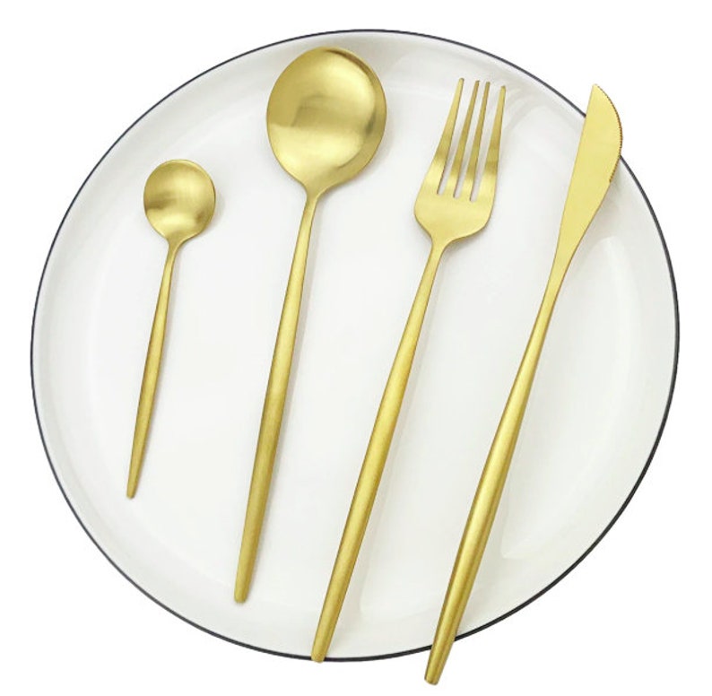 May include: A set of four gold-colored utensils: a small spoon, a large spoon, a fork, and a knife. The utensils are arranged on a white plate with a black rim. The utensils have a sleek, modern design.