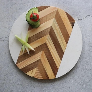 May include: A round serving board with a chevron pattern made of wood and marble. The wooden section features various shades of brown. A slice of cucumber with a cherry tomato and green onion garnish are on the board.