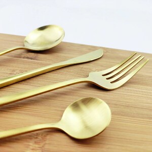 May include: A close-up shot of gold-colored flatware on a wooden surface. The set includes a spoon, knife, and fork. The spoon has a rounded bowl, the knife has a rounded tip, and the fork has four tines. The flatware has a matte finish.
