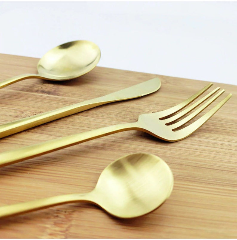May include: A close-up shot of gold-colored flatware on a wooden surface. The set includes a spoon, knife, and fork. The spoon has a rounded bowl, the knife has a rounded tip, and the fork has four tines. The flatware has a matte finish.