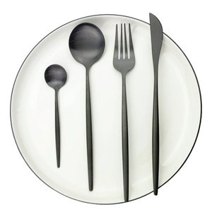 May include: A set of black matte finish silverware on a white plate with a black rim. The set includes a small spoon, a large spoon, a fork, and a knife. The plate is round and the silverware is arranged neatly.