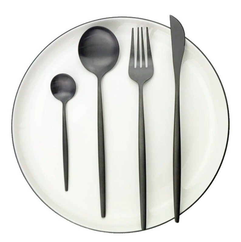 May include: A set of black matte finish silverware on a white plate with a black rim. The set includes a small spoon, a large spoon, a fork, and a knife. The plate is round and the silverware is arranged neatly.