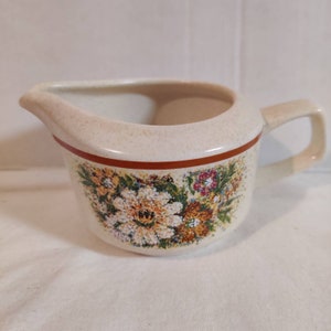 May include: A white ceramic creamer with a brown rim and a floral design. The flowers are yellow, orange, and pink.