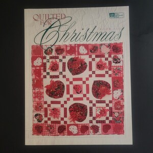 Könnte beinhalten: Ein Buch mit dem Titel "Quilted for Christmas" mit einem roten und weißen Quiltmuster auf dem Cover. Der Quilt zeigt ein Muster aus Herzen und Quadraten mit roten Weihnachtsstern-Designs.