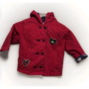 May include: A red hooded jacket with a double-breasted button closure. The jacket features two heart-shaped patches, one with a striped design and the other with a green heart on a navy background. The cuffs have a patterned design.