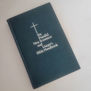 Puede incluir: Un libro de tapa dura de color azul oscuro con una cruz blanca y el título "The Parallel New Testament and Unger's Bible Handbook".
