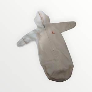 May include: A cream-colored baby sleep sack with a hood and long sleeves. The sleep sack has a full-length zipper and an embroidered orange dinosaur on the chest. The soft, plush material is designed for comfort and warmth.