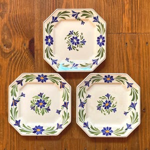 May include: Three octagonal ceramic plates with a floral design. Each plate features a central cluster of blue flowers and a border of blue flowers and green leaves on a cream background. The plates are arranged on a wooden surface.
