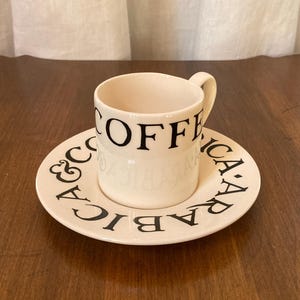 May include: A white ceramic coffee cup and saucer set. The cup has the word "COFFEE" in black lettering. The saucer is also white with black text that reads "ARABICA & COFFEE". The set is on a wooden surface.