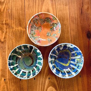 May include: Three small, round ceramic bowls with colorful, abstract designs. One bowl is orange and green, another is blue and white, and the third is green and blue. The bowls are set on a wooden surface.