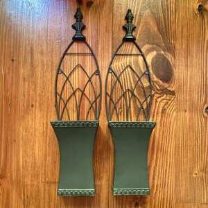 May include: A pair of dark green metal wall planters featuring a solid rectangular base with a decorative beaded border and an arched wire trellis top topped with a pointed finial. The planters have a smooth finish and a symmetrical design.