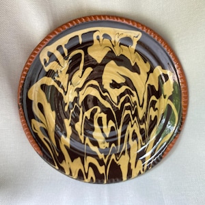 May include: A handmade ceramic plate with a brown and yellow swirled design. The plate has a dark brown center with a textured, light brown rim. The yellow pattern creates a marbled effect, adding visual interest to the piece.