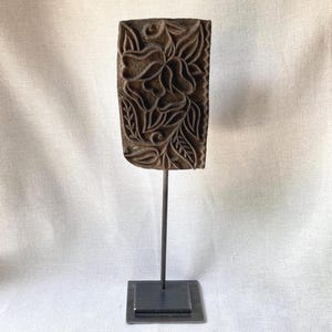 May include: A rectangular dark brown wooden block with a carved floral pattern featuring petals and leaves. The block is mounted vertically on a thin black metal rod attached to a square black metal base, creating a standing display piece.