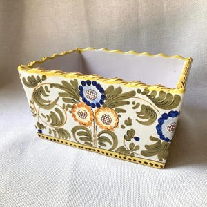May include: Rectangular ceramic planter with a scalloped yellow rim. The planter is decorated with floral and leaf patterns in blue, orange, and green on a cream background. A yellow border with brown dots accents the bottom.