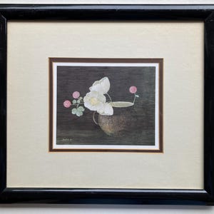 May include: Framed floral still life art print. The artwork features white flowers and pink buds in a brown vase against a dark background. The print is framed in a black wooden frame with a cream-colored mat.