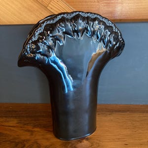 May include: A glossy black ceramic vase with a unique, flared top. The vase has a tall, cylindrical body that widens towards the top, which features a decorative, scalloped edge. The vase is displayed on a wooden surface.