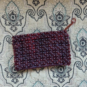 Op de afbeelding: Een rechthoekige, met kralen bezette clutch in tinten bordeauxrood en rood. De kralen zijn dicht op elkaar gepakt, waardoor een textuur ontstaat. Een kleine lus is bevestigd. De clutch ligt op een achtergrond met patroon.