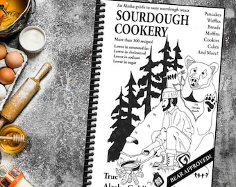 May include: A spiral-bound cookbook titled "SOURDOUGH COOKERY" with a black and white illustration of a man cooking over a fire and a bear. The cover text includes "An Alaska guide to tasty sourdough treats" and "True Alaska Gold!"