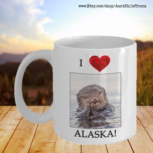 May include: White ceramic coffee mug with a red heart and the text "I love Alaska!"  A photo of a sea otter in the water is in the center of the mug.