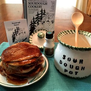 May include: A white ceramic sourdough pot with green trim and the text "Sour Dough Pot" on the side. A wooden spoon is inside the pot. A stack of pancakes with syrup on a white plate with green trim. A small bottle of maple syrup and a book titled "Sourdough Cookery" are in the background.