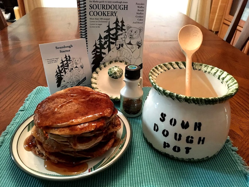 May include: A white ceramic sourdough pot with green trim and the text "Sour Dough Pot" on the side. A wooden spoon is inside the pot. A stack of pancakes with syrup on a white plate with green trim. A small bottle of maple syrup and a book titled "Sourdough Cookery" are in the background.