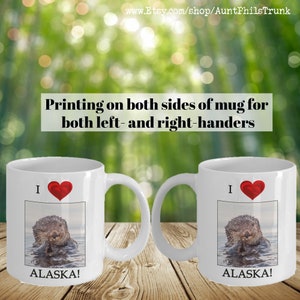 May include: White ceramic mug with a red heart and the text "I  ALASKA!" printed on it. The mug features a photo of a sea otter in the water. The mug is printed on both sides.