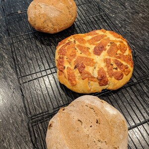 May include: Three loaves of freshly baked bread on a wire rack. The bread on the left and right are round and have a rustic appearance. The bread in the middle is round and has a golden brown crust with a pattern of small holes.