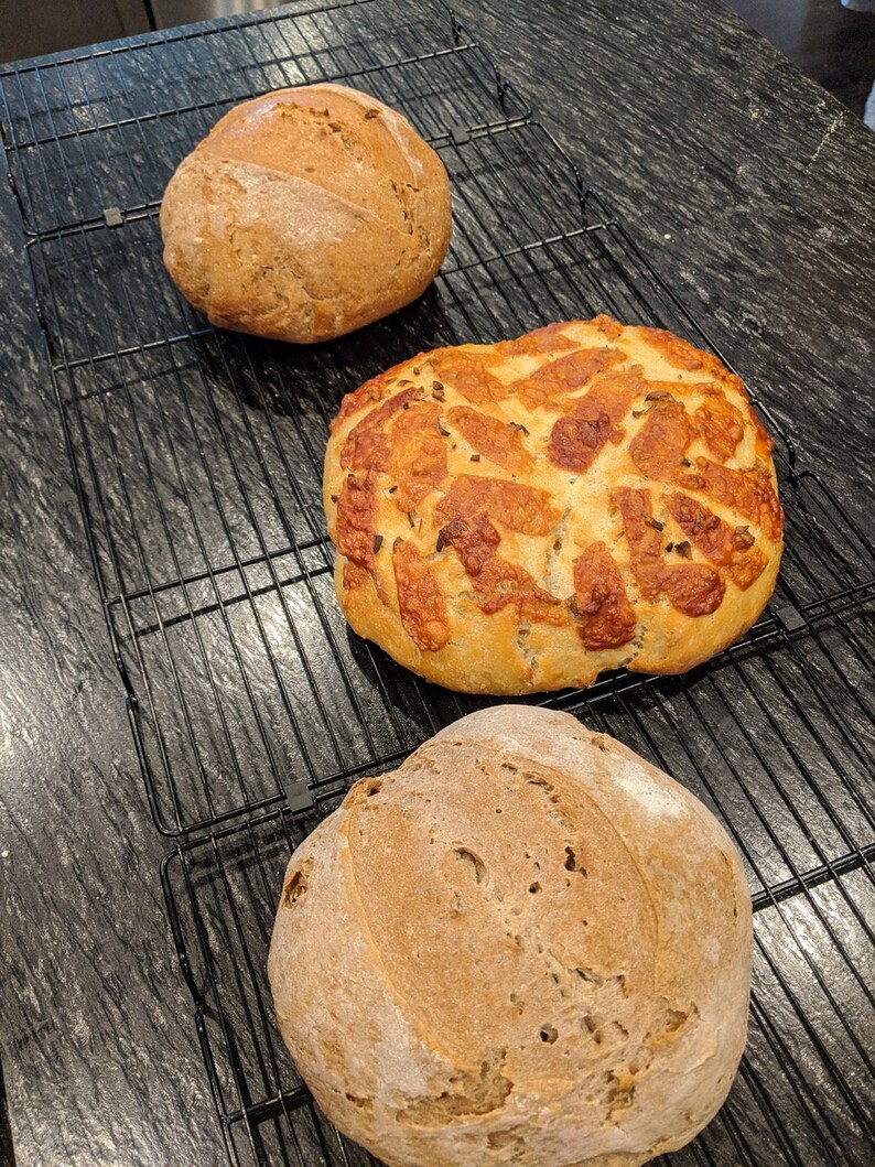 May include: Three loaves of freshly baked bread on a wire rack. The bread on the left and right are round and have a rustic appearance. The bread in the middle is round and has a golden brown crust with a pattern of small holes.