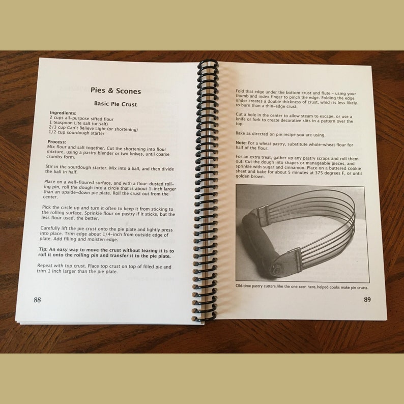 May include: Open cookbook with spiral binding, displaying a recipe for "Pies & Scones" and "Basic Pie Crust." The pages show instructions and a photo of a pastry cutter. The book is open on a wooden surface.