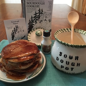 May include: A white ceramic sourdough pot with green trim and the words "Sour Dough Pot" written on it. A wooden spoon is inside the pot. A stack of pancakes with syrup on a plate, a bottle of maple syrup, and a sourdough starter booklet are also in the image.