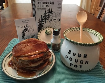 May include: A white ceramic sourdough pot with green trim and the words "Sour Dough Pot" written on it. A wooden spoon is inside the pot. A stack of pancakes with syrup on a plate, a bottle of maple syrup, and a sourdough starter booklet are also in the image.