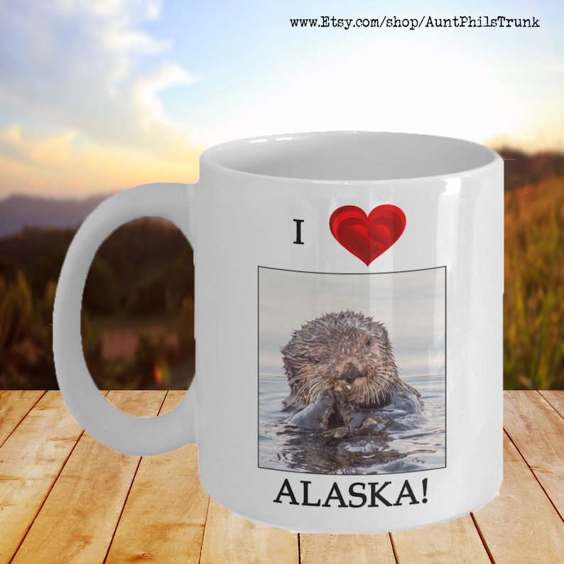 May include: White ceramic coffee mug with a red heart and the text "I love Alaska!"  A photo of a sea otter in the water is in the center of the mug.