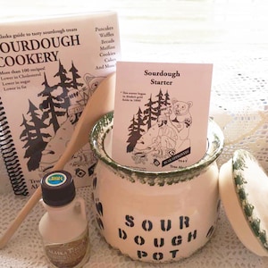 May include: A sourdough starter kit with a white ceramic sourdough pot with a green lid, a wooden spoon, a small bottle of maple syrup, a sourdough starter packet, and a cookbook titled "Sourdough Cookery".