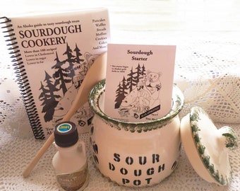 May include: A sourdough starter kit with a white ceramic sourdough pot with a green lid, a wooden spoon, a small bottle of maple syrup, a sourdough starter packet, and a cookbook titled "Sourdough Cookery".
