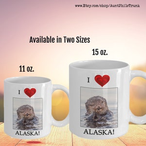 May include: White ceramic mug with a red heart and the text "I love Alaska!"  The mug features a photo of a sea otter in the water. The mug is available in two sizes: 11 oz and 15 oz.