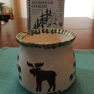 May include: A white ceramic mug with a green rim and a moose silhouette design. The mug is sitting on a blue placemat.