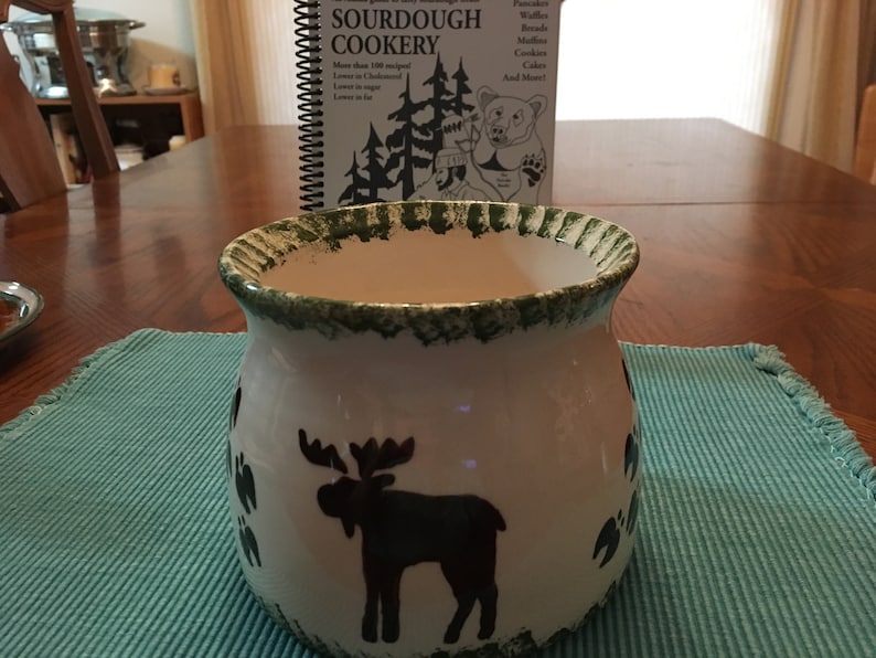 May include: A white ceramic mug with a green rim and a moose silhouette design. The mug is sitting on a blue placemat.