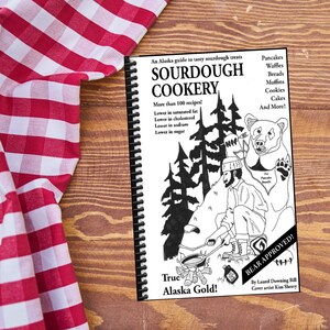 May include: A spiral-bound cookbook titled "SOURDOUGH COOKERY" with a black and white illustration of a man cooking over a fire, a bear, and trees. The cover text includes "True Alaska Gold!" and "BEAR APPROVED!" The book is on a wooden surface next to a red and white checkered cloth.
