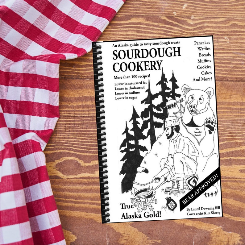 May include: A spiral-bound cookbook titled "SOURDOUGH COOKERY" with a black and white illustration of a man cooking over a fire, a bear, and trees. The cover text includes "True Alaska Gold!" and "BEAR APPROVED!" The book is on a wooden surface next to a red and white checkered cloth.