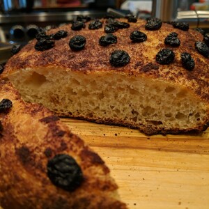 May include: A loaf of freshly baked bread with a golden-brown crust, topped with dark, dried fruit. A slice has been cut, revealing the airy interior. The bread rests on a wooden cutting board.