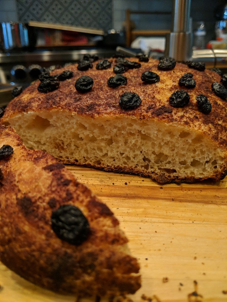 May include: A loaf of freshly baked bread with a golden-brown crust, topped with dark, dried fruit. A slice has been cut, revealing the airy interior. The bread rests on a wooden cutting board.