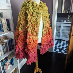 May include: A cape made of fall leaves in shades of green, yellow, orange, and red. The leaves are arranged in a gradient pattern, starting with green at the top and transitioning to red at the bottom. The cape is displayed on a mannequin.