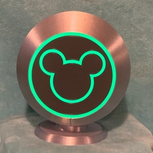 May include: A silver-colored, circular decorative object with a green glowing outline of a Mickey Mouse head. The object has a dark brown center and sits on a small, matching silver base. The design is simple and modern.