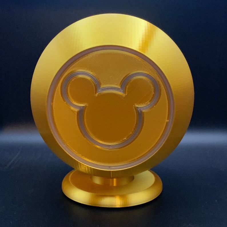 May include: A gold-colored, round decorative object featuring a Mickey Mouse silhouette in the center. The object has a circular base and a clear, translucent ring around the Mickey Mouse design. The overall design is smooth and shiny.