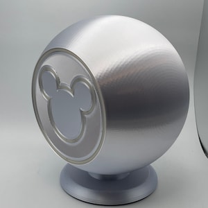 May include: A silver sphere-shaped decorative object with a Mickey Mouse silhouette on the front. The object is mounted on a circular base. The design features a textured surface and a smooth, metallic finish.